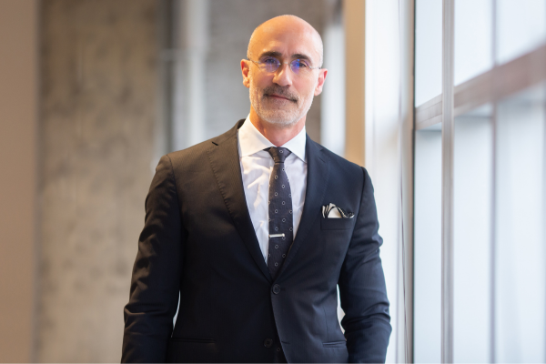 Arthur Brooks Professional Portrait, Happiness Expert, Bestselling Author, Harvard Professor, And Keynote Speaker, Standing In A Tailored Navy Suit Near A Window In A Modern Business Setting.