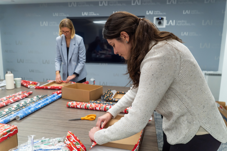 Two Women Wrap Holiday Gifts At A Large Conference Table Covered With Rolls Of Festive Wrapping Paper, Scissors, And Boxes. One Woman In A Light Sweater Focuses On Folding Paper Around A Box In The Foreground, While Another In A Light Blazer Works On A Gift In The Background. A Screen And A Wall With The Leading Authorities Logo Are Behind Them.