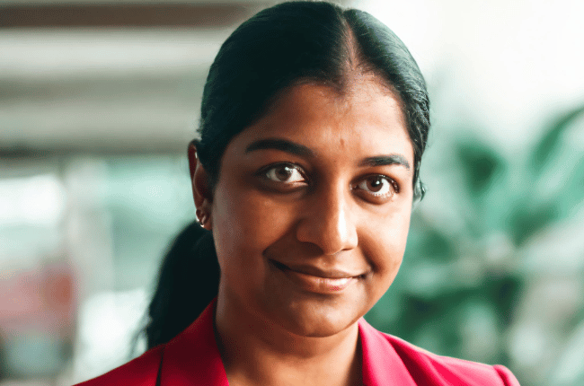 Priyanka Rao, a professional speaker, wearing a bright red blazer over a white top, standing in an elegant indoor setting with a blurred green plant in the background, exuding confidence and approachability